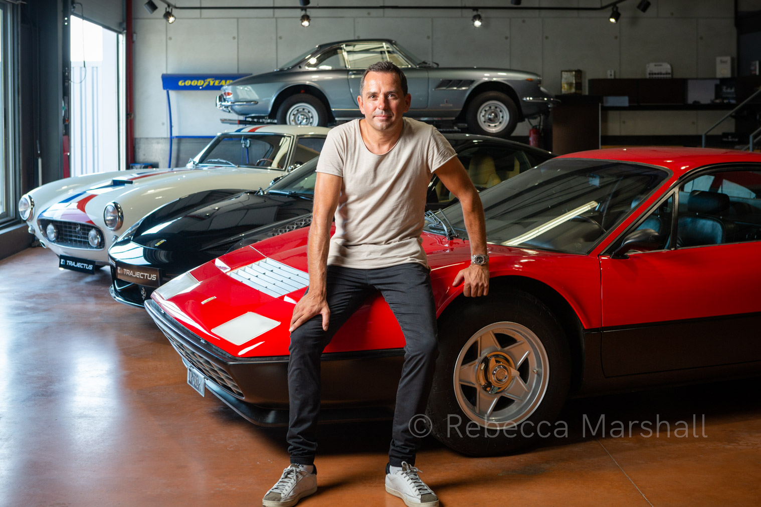 Man poses for the photographer sitting on the bonnet of his red vintage Ferrari in a garage with other vintage cars in the background