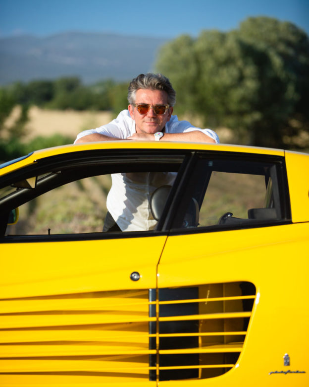 Man in white shirt poses for the photographer leaning on the roof of his yellow Ferrari