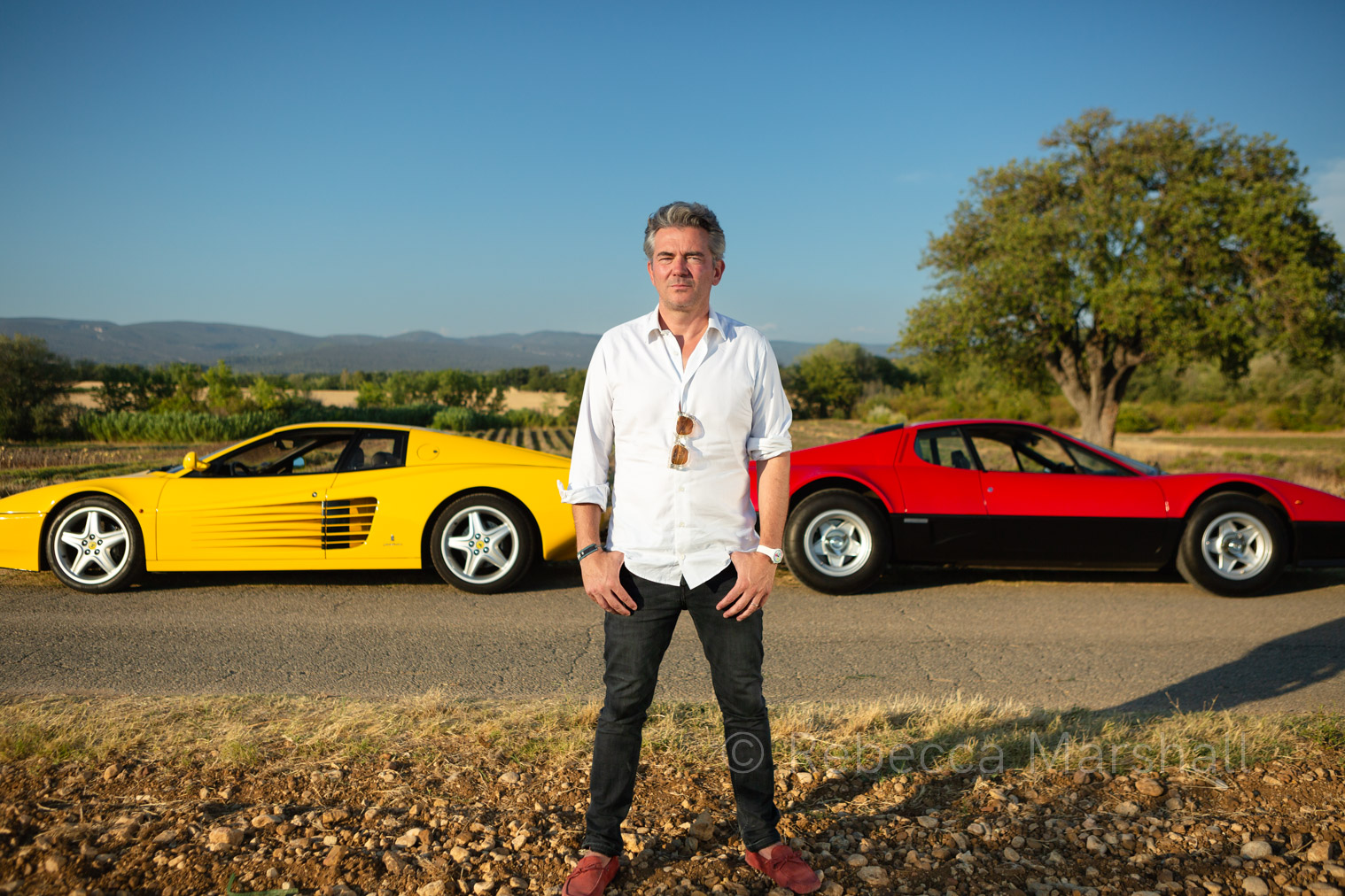 Man in white shirt poses for the photographer in front of a yellow and a red Ferrari