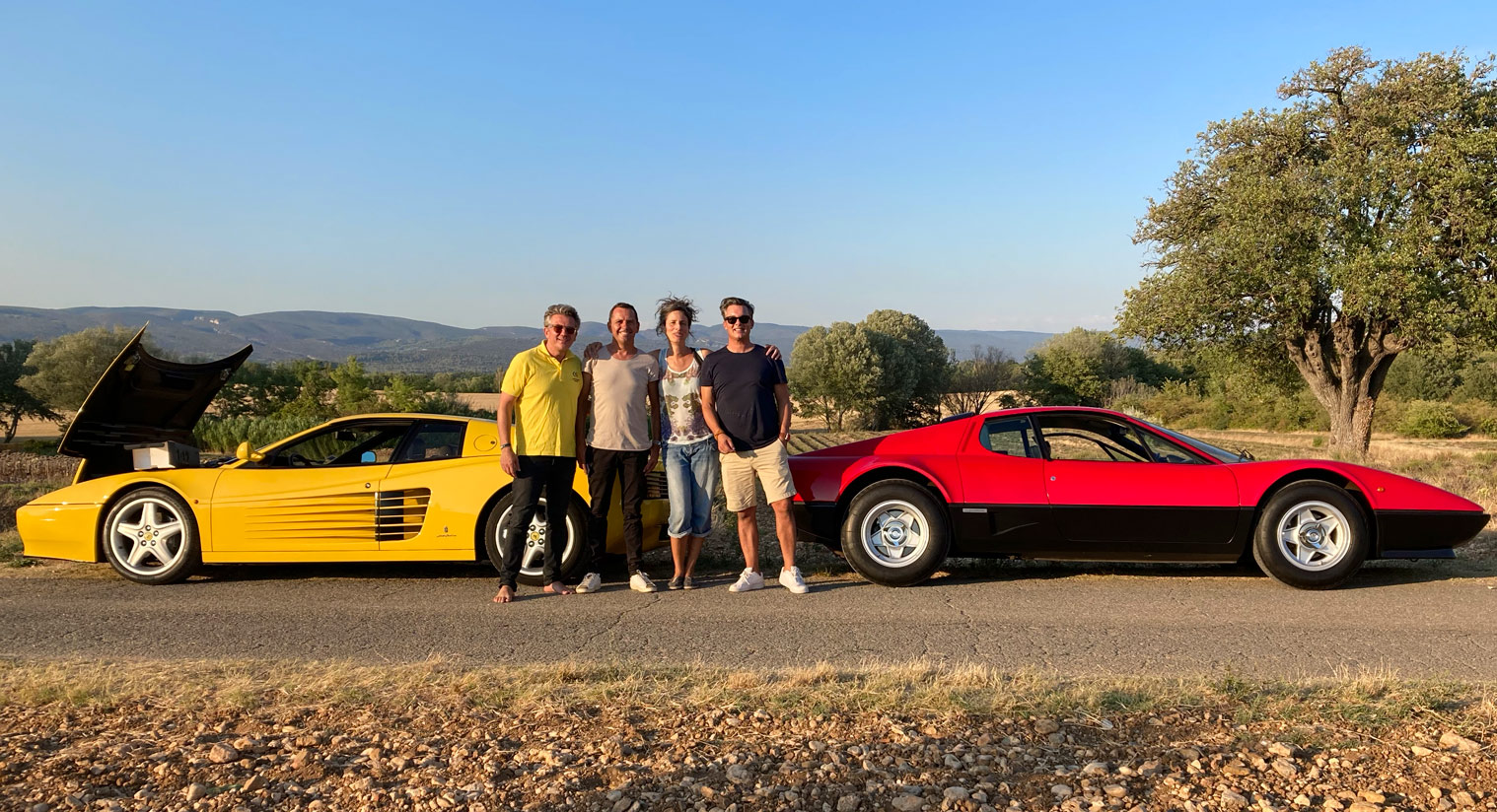 Photo of 4 people posing for the camera on a roadside between yellow and red Ferrari cars