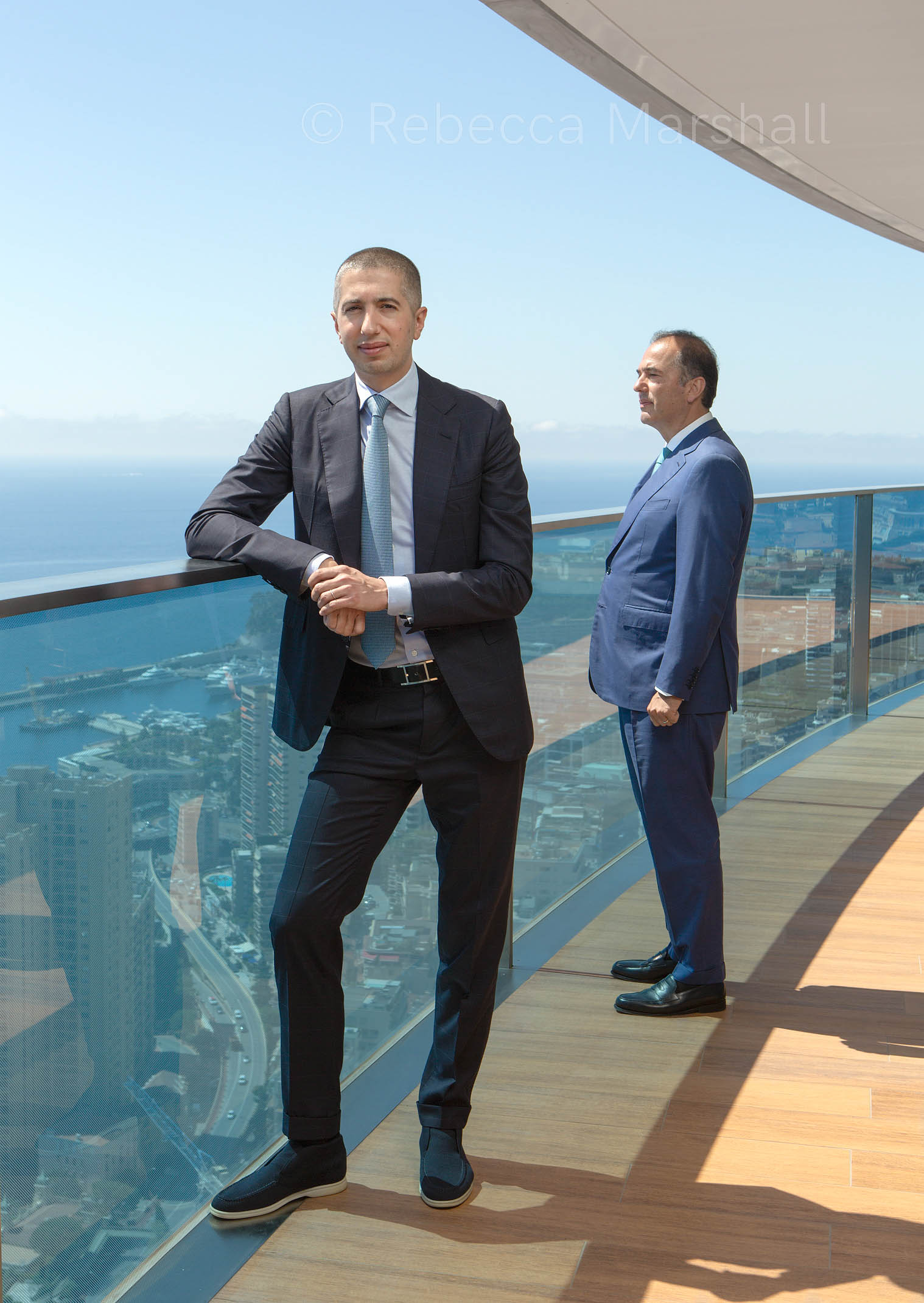 Two men in suits stand on the balcony of an upper-floor apartment, a view of Monaco beneath them