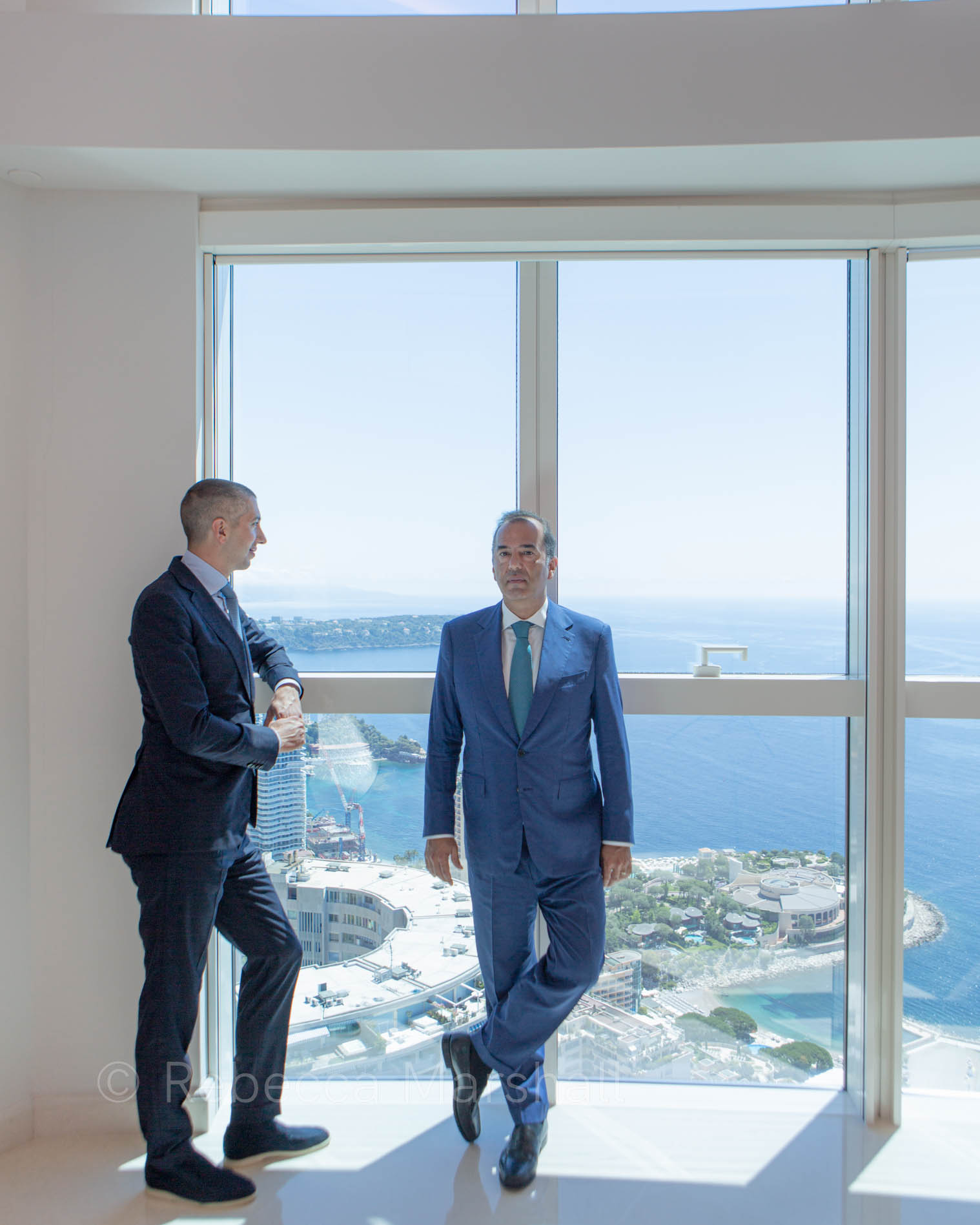 Two men in suits stand by the large window of an upper-floor apartment, a view of Monte Carlo beneath them