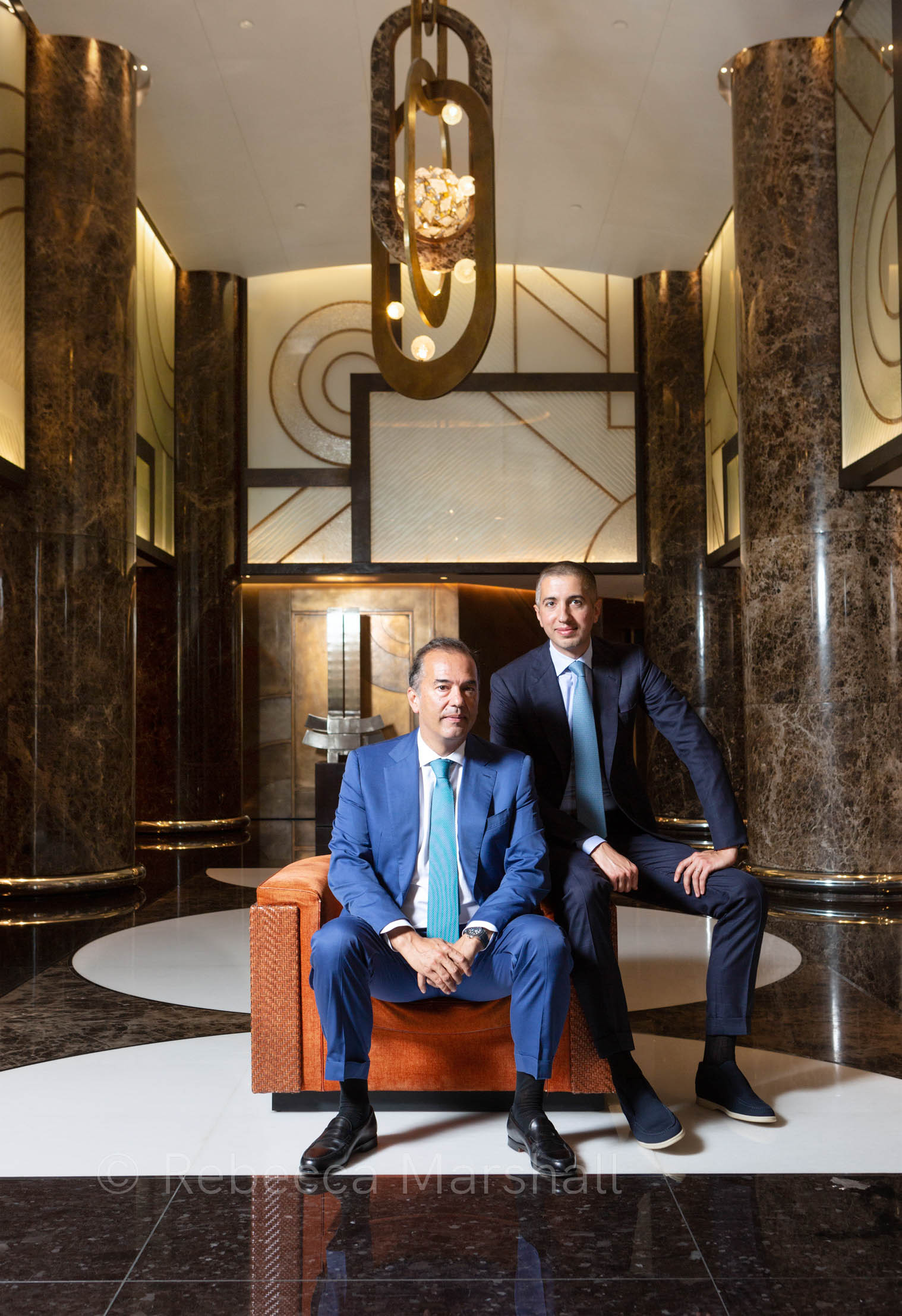 Two men in suits sit on an orange armchair in the foyer of a luxury residence