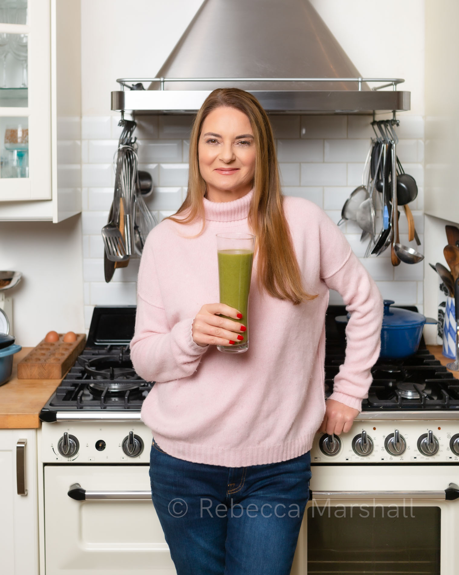 Photograph of a woman in a pink jumper in a kitchen, holding a green smoothie