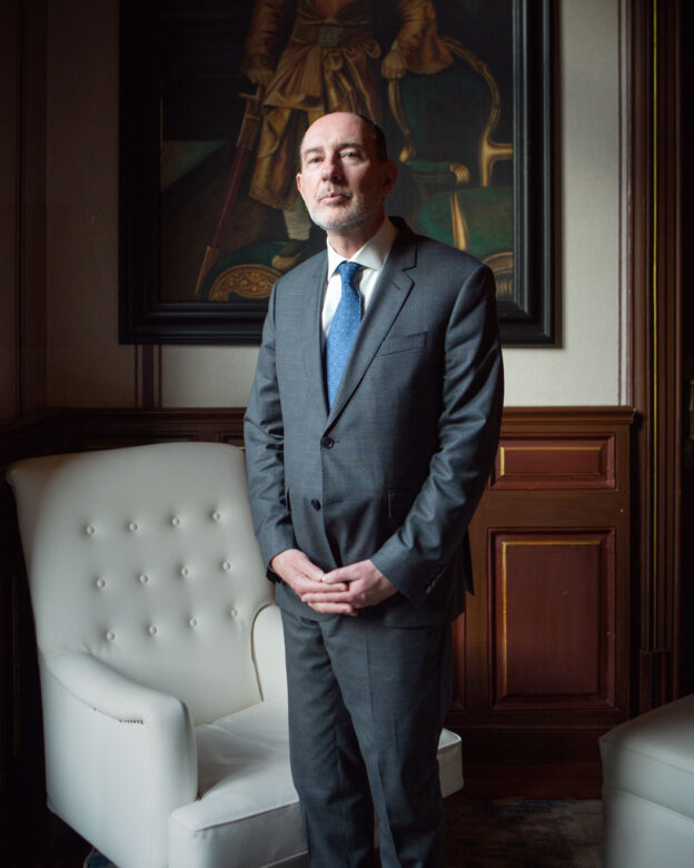 Portrait of Pierre De Gaulle, dressed in a suit and blue tie, standing in a formal salon