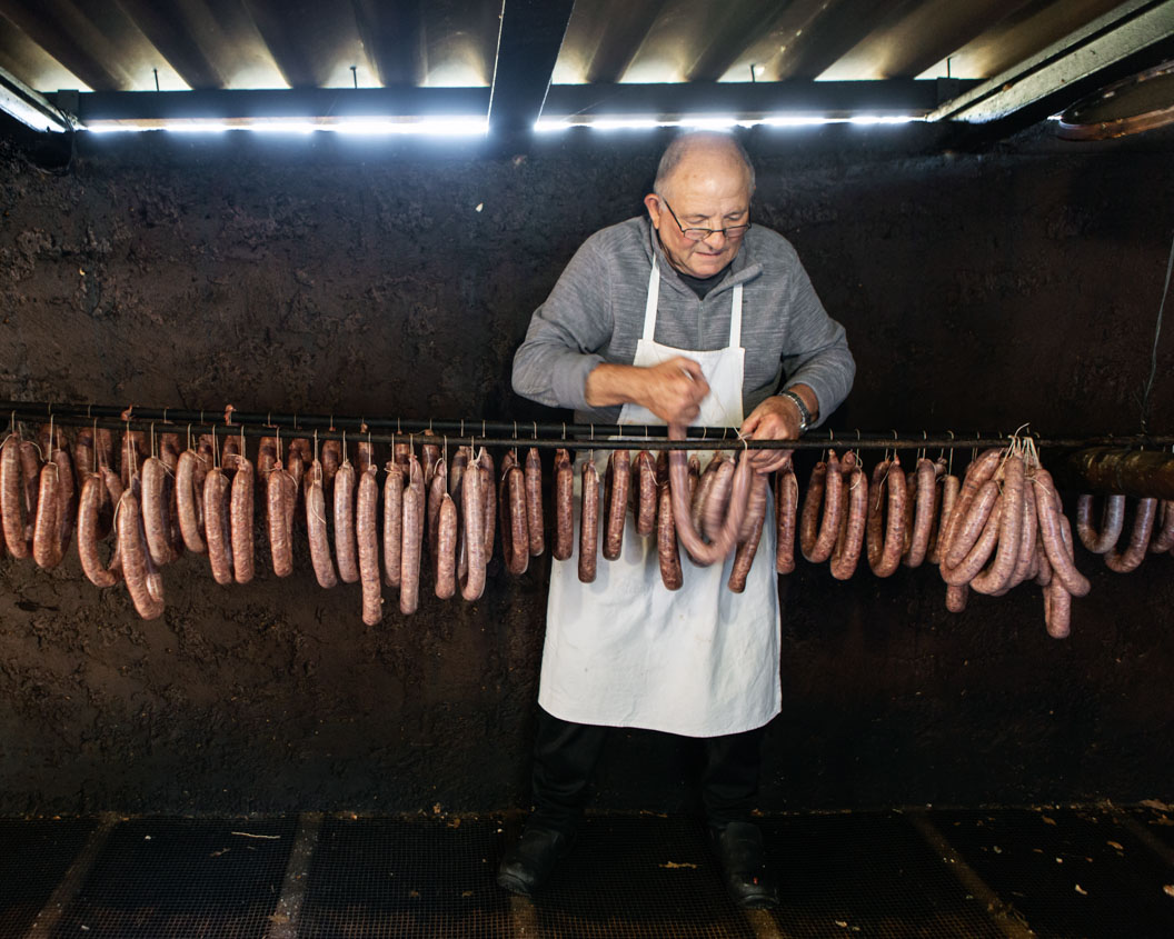 An elderly man strings up a line of sausages to cure in a dark shed